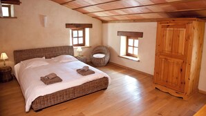A bedroom with a large bed, two nightstands, a wicker chair, a wooden wardrobe, and two windows. - Gîte Petite Maison in the Gers countryside (Castelnau-sur-l'Auvignon)