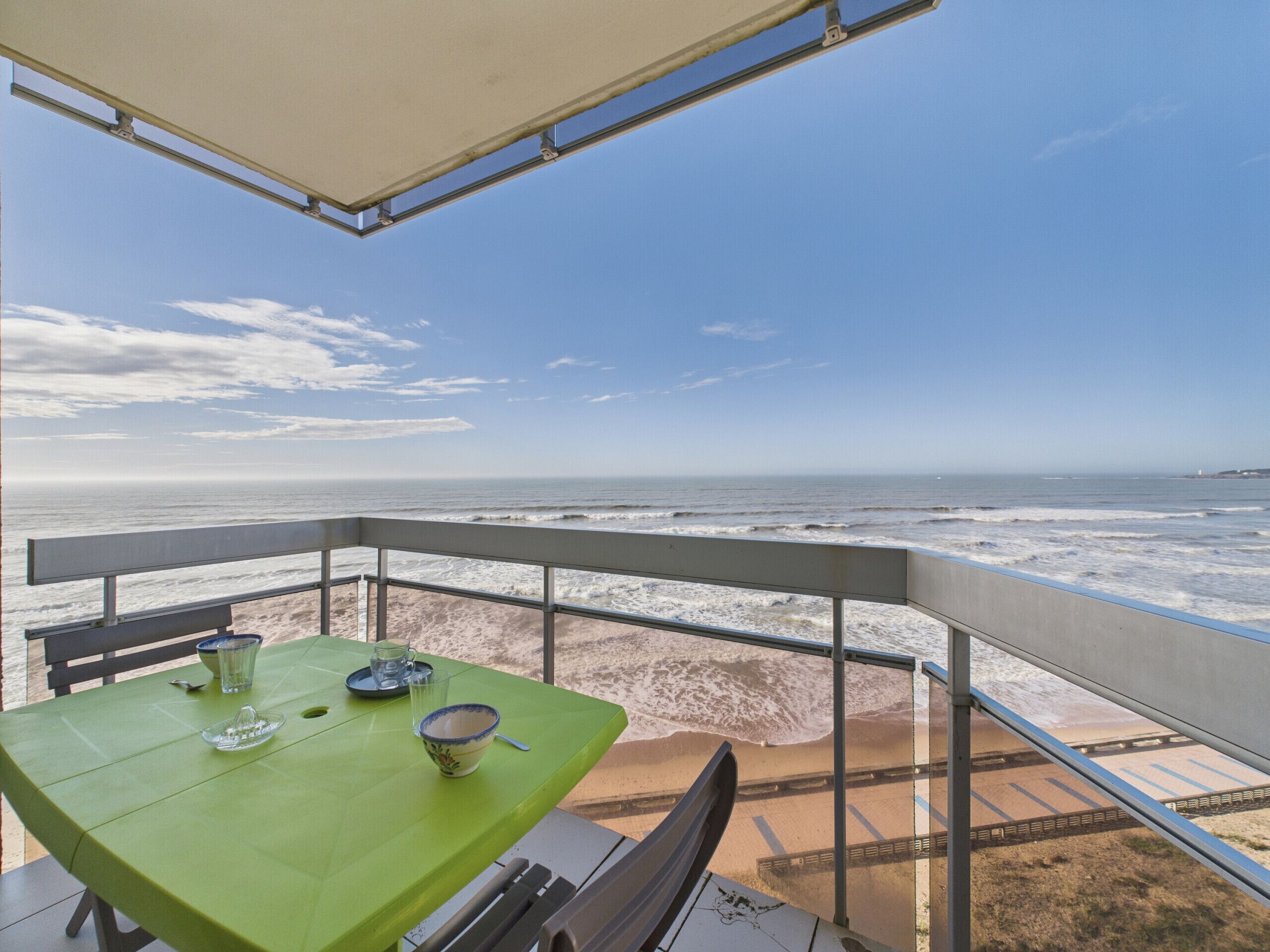 A balcony with a table and chairs, overlooking a beach with waves and a clear sky.
