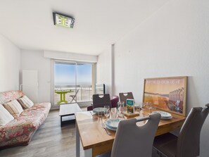 A modern hotel room with a balcony, a dining table set with glasses and plates, a floral sofa, and a painting of Saint Gilles Croix de Vie. - Accommodation for 4 with magnificent sea view (Saint-Gilles-Croix-de-Vie)