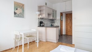 Cocina pequeña con una mesa y sillas blancas, una cafetera y una puerta de madera que da a otra habitación.