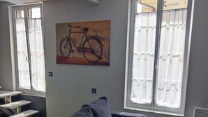 Un salon moderne avec un canapé gris, un tableau représentant un vélo et de grandes fenêtres avec des rideaux.