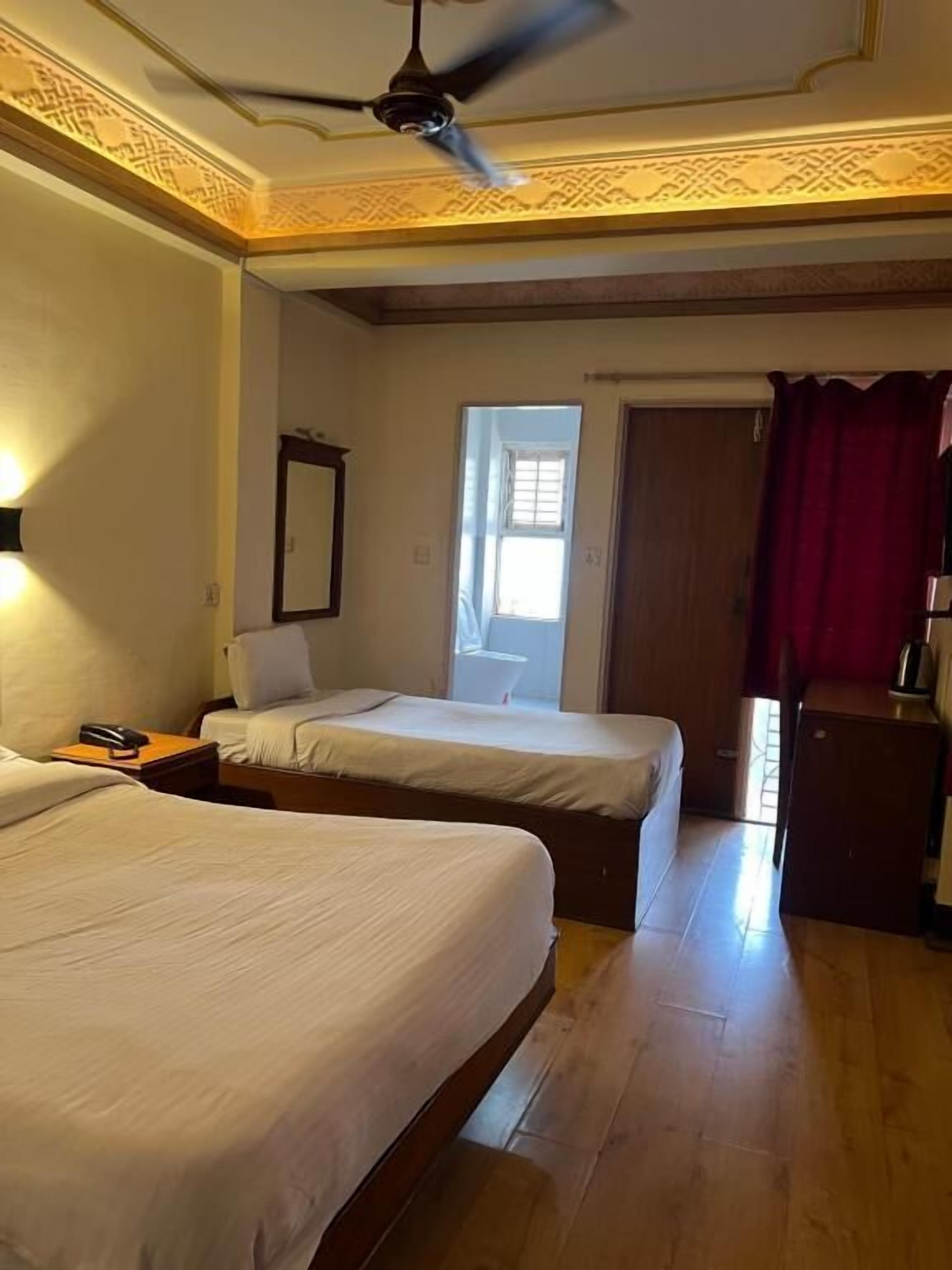 A hotel room with two beds, a ceiling fan, a window, a mirror, and a small desk with a lamp.