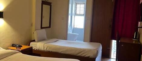Quarto de hotel com duas camas, um ventilador de teto, uma janela, um espelho e uma escrivaninha pequena com abajur.