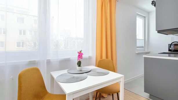 Una cocina moderna con encimera blanca, un lavamanos y un ventanal con vistas a edificios. Hay una mesa con dos sillas y una pequeña planta en maceta.