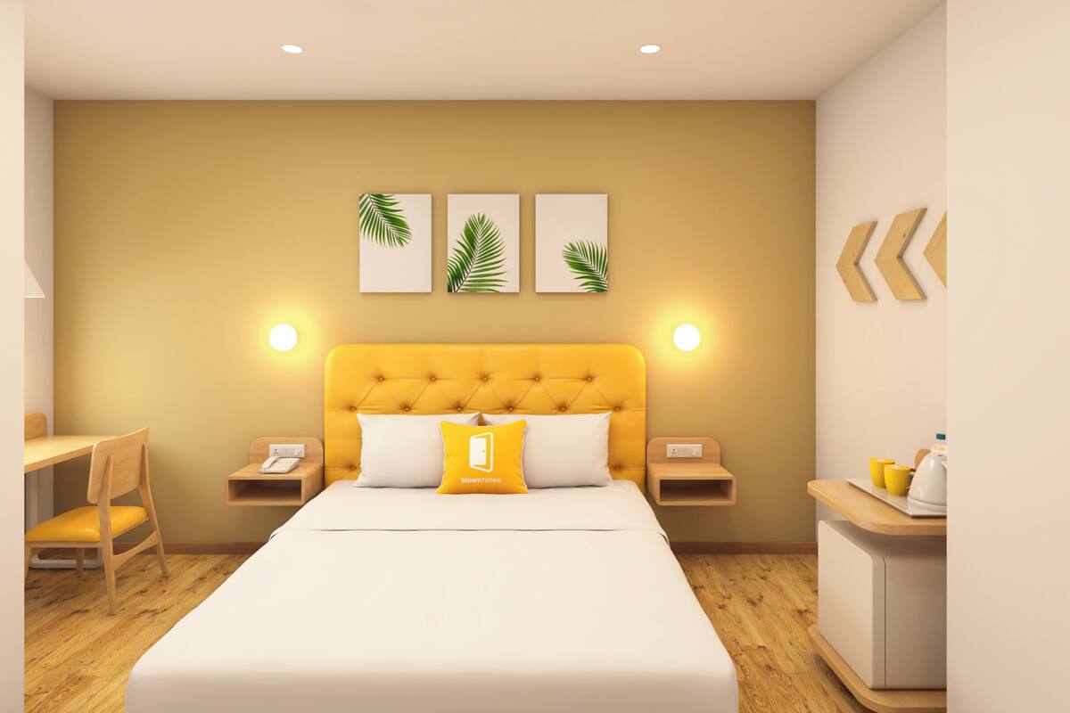 A modern hotel room with a yellow tufted headboard, a bed with white and yellow bedding, a desk with a chair, and a bedside table with a lamp.