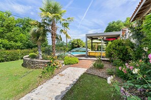 Un sentier de jardin mène à un espace de repos abrité, avec une piscine en arrière-plan.