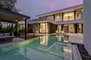 Een modern huis met twee verdiepingen, een slaapkamer met glazen wanden, een zwembad en een patio met buitenmeubilair.