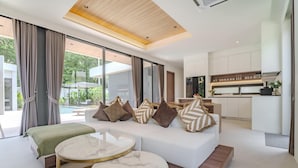 A modern living room with a white sofa, two marble-topped coffee tables, and a view of a pool and garden through large windows. - Aileen Layan Villa 6/ 4BR Forest/ 2 Minutes Beach (Choeng Thale)