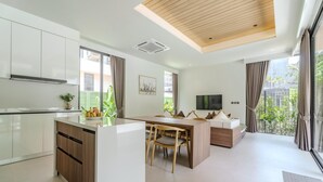 A modern kitchen with a central island, a dining area with chairs, a sofa, and a TV. - Aileen Layan Villa 6/ 4BR Forest/ 2 Minutes Beach (Choeng Thale)