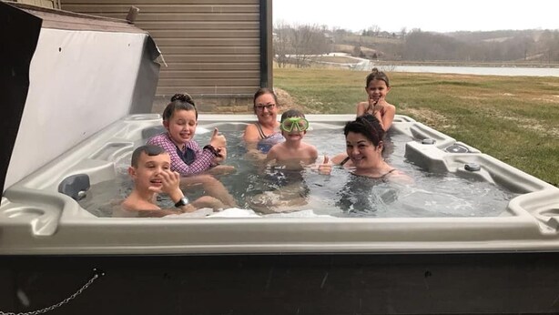 En gruppe mennesker i en jacuzzi med udsigt til en sø og en bygning i baggrunden.