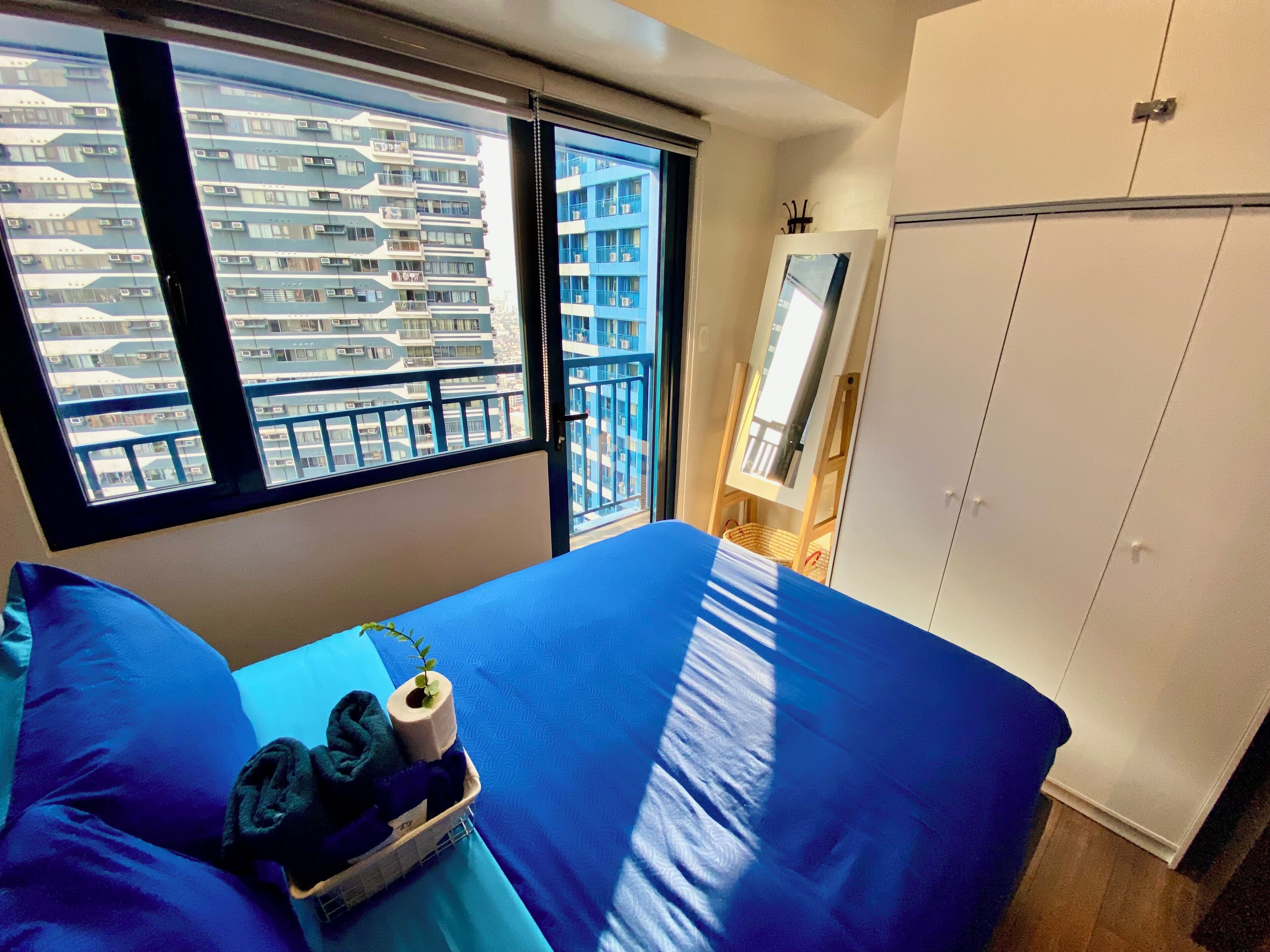 A bedroom with a large bed, a window with a view of a building, a wardrobe, and a mirror on the wall.