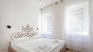 Una cama bien hecha con sábanas blancas y dos toallas enrolladas, un cabecero de metal decorativo, y una ventana con cortinas translúcidas.