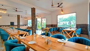 En restaurang med långa träbord och blå klädda stolar, stenkylor och stora fönster som ger utsikt över grönområden.
