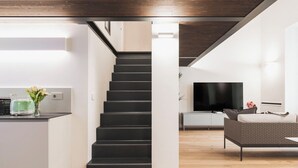 Ein moderner Wohnraum mit einer Treppe, die zu einer zweiten Etage führt, einer Küchenzeile mit Toaster und Blumen, einem Flachbildfernseher und einem grauen Sofa.
