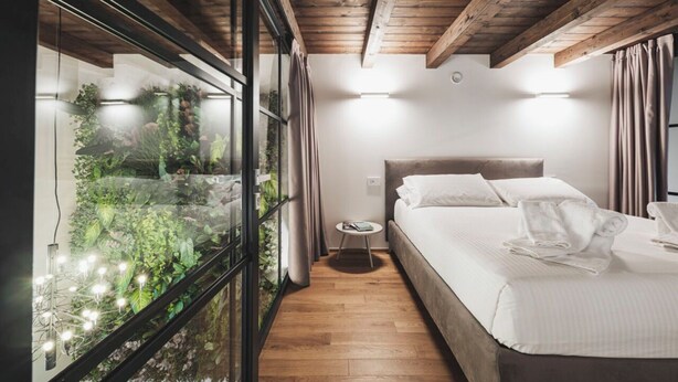 Ein Schlafzimmer mit Bett, Nachttisch und Blick auf einen Garten durch eine Glaswand.