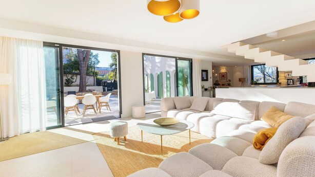 Ein modernes Wohnzimmer mit einer großen Couch, einem Couchtisch und einem Essbereich mit Stühlen. Eine Treppe führt zu einer oberen Ebene.