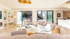 Ein modernes Wohnzimmer mit einer großen Couch, einem Couchtisch und einem Essbereich mit Stühlen. Links befindet sich ein Bücherregal und ein Fernseher.