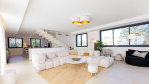 Ein großzügiger Wohnbereich mit einem weißen Modulsofa, einem runden Couchtisch und einem schwarzen Sofa. Große Fenster sorgen für natürliches Licht und bieten einen Blick ins Freie.