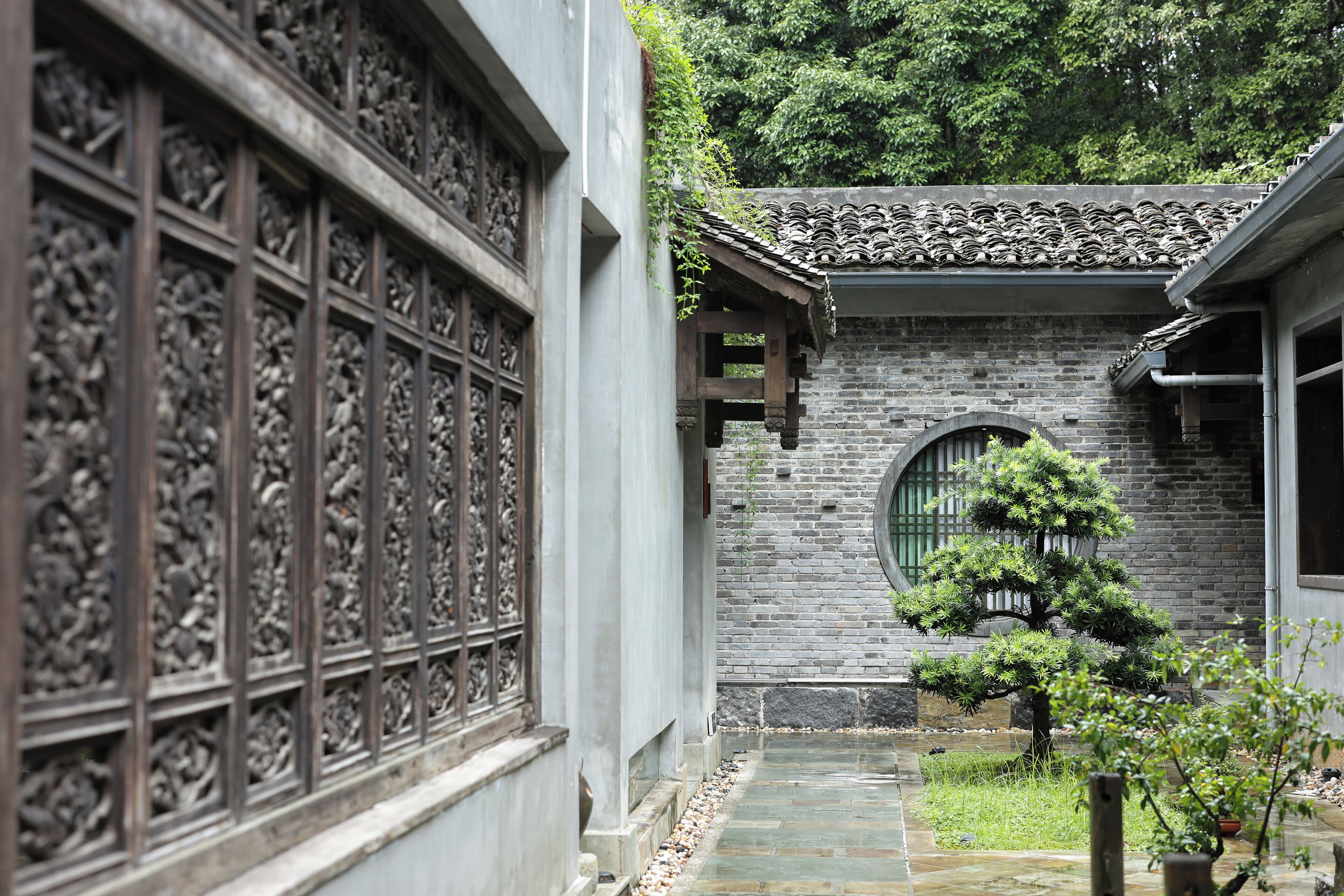 A traditional courtyard with a stone pathway, a bonsai tree, and a building with intricate window designs.