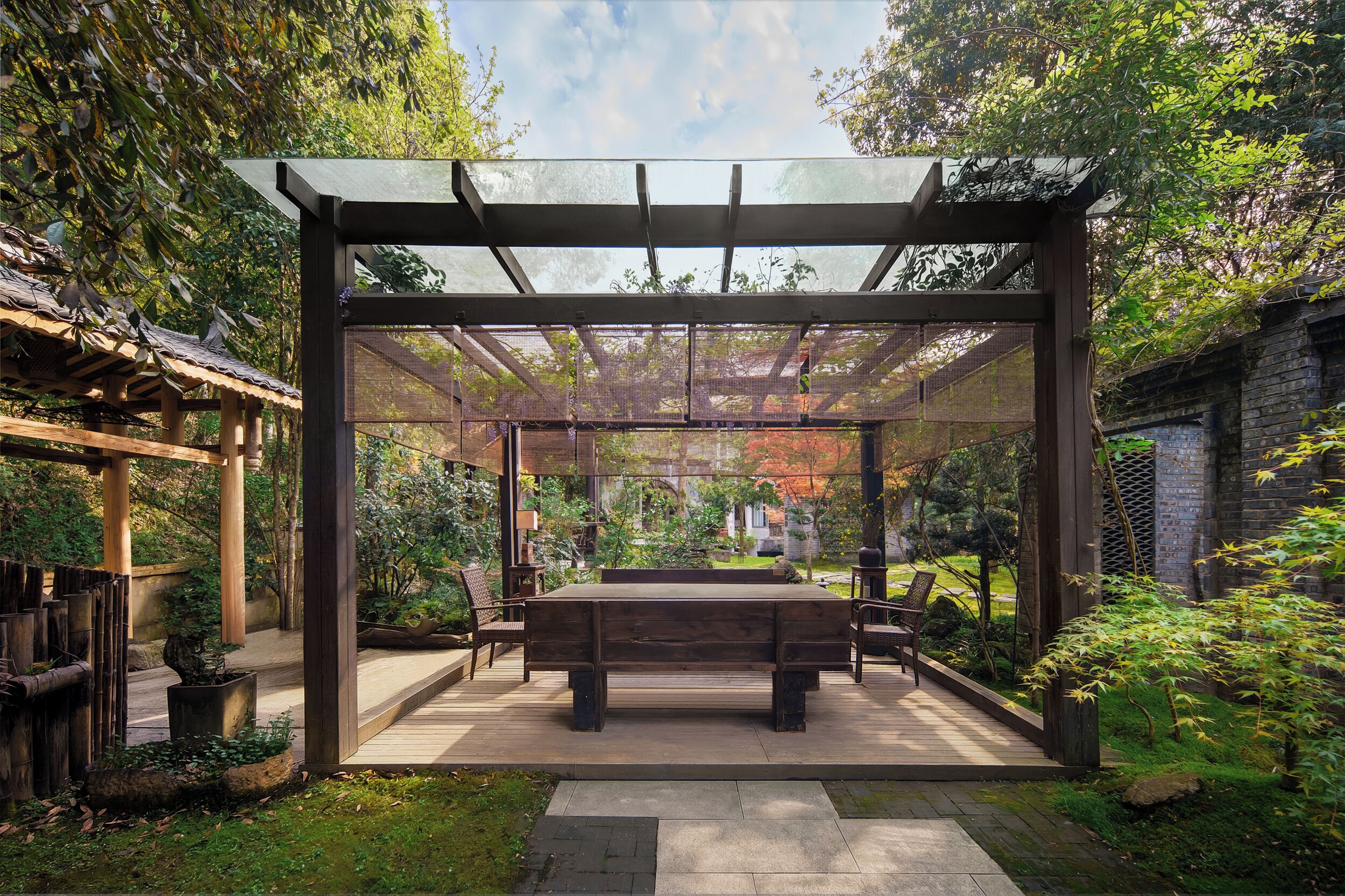 Un espacio al aire libre cubierto con mesa y sillas, rodeado de follaje y una estructura de pérgola.