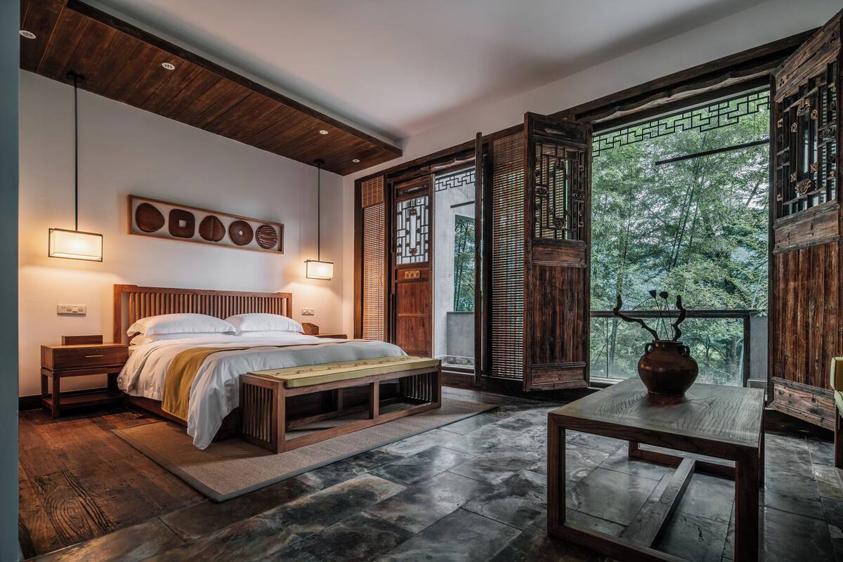 Ein Schlafzimmer mit einem hölzernen Bett, Nachttischen, einer Bank, einem Tisch und Blick ins Grüne durch ein großes Fenster.