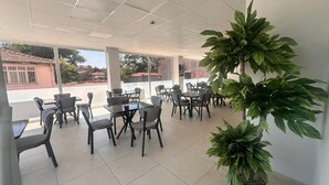 Un espace intérieur vaste et lumineux, avec de nombreuses tables et chaises, de grandes fenêtres et des plantes en pot.