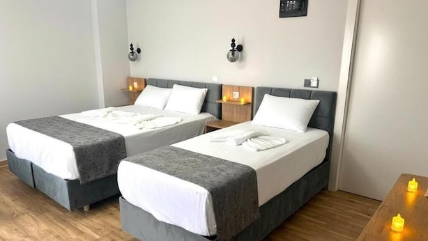 Une chambre d’hôtel avec deux lits, chacun doté d’une tête de lit grise et de linge de lit blanc. On y trouve des tables de chevet avec des lampes, ainsi qu’une petite étagère également équipée d’une lampe. La chambre est recouverte de parquet et comprend une table en bois ornée de bougies.