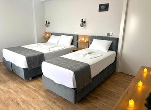 A hotel room with two beds, each with a grey headboard and white bedding. There are bedside tables with lamps and a small shelf with a lamp. The room has wooden flooring and a wooden table with candles. - AVKA PALACE (Edirne)