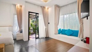 Une chambre d’hôtel avec un lit, une fenêtre agrémentée de coussins bleus, une porte donnant sur l’extérieur et une vue sur la verdure.