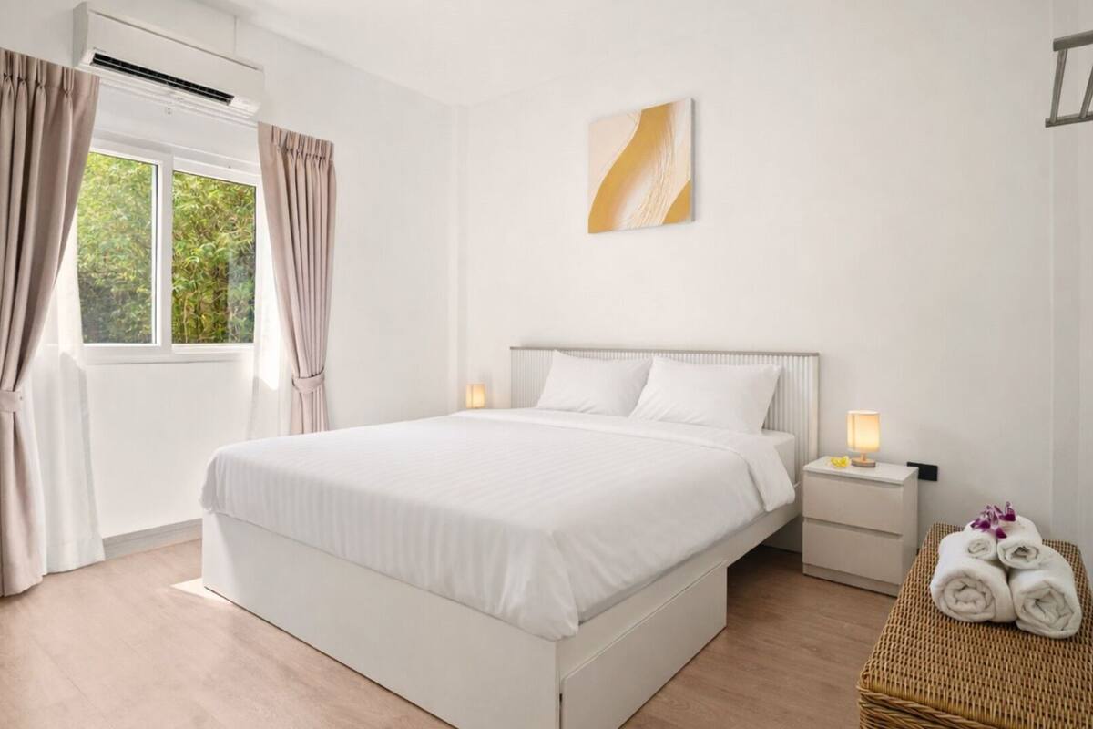 A hotel room with a bed, bedside tables, a window with curtains, a wall-mounted air conditioner, and a painting.