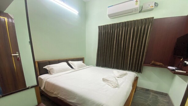 A hotel room with a bed, a wooden headboard, a bedside table, a wall-mounted air conditioner, and a window with curtains. - MGM Suite Rooms (Tiruvannamalai)