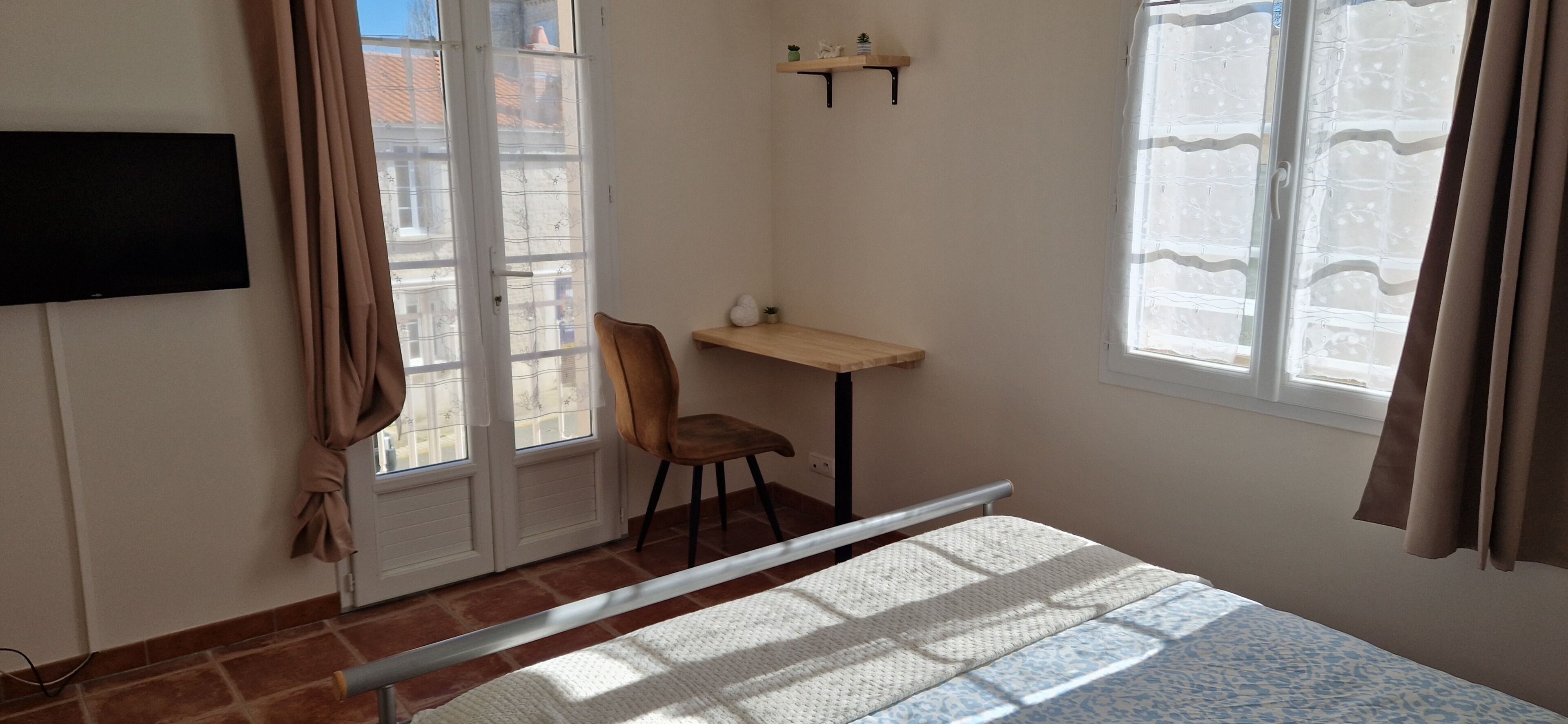 A bedroom with a bed, a desk, a chair, a TV mounted on the wall, and a window with curtains.