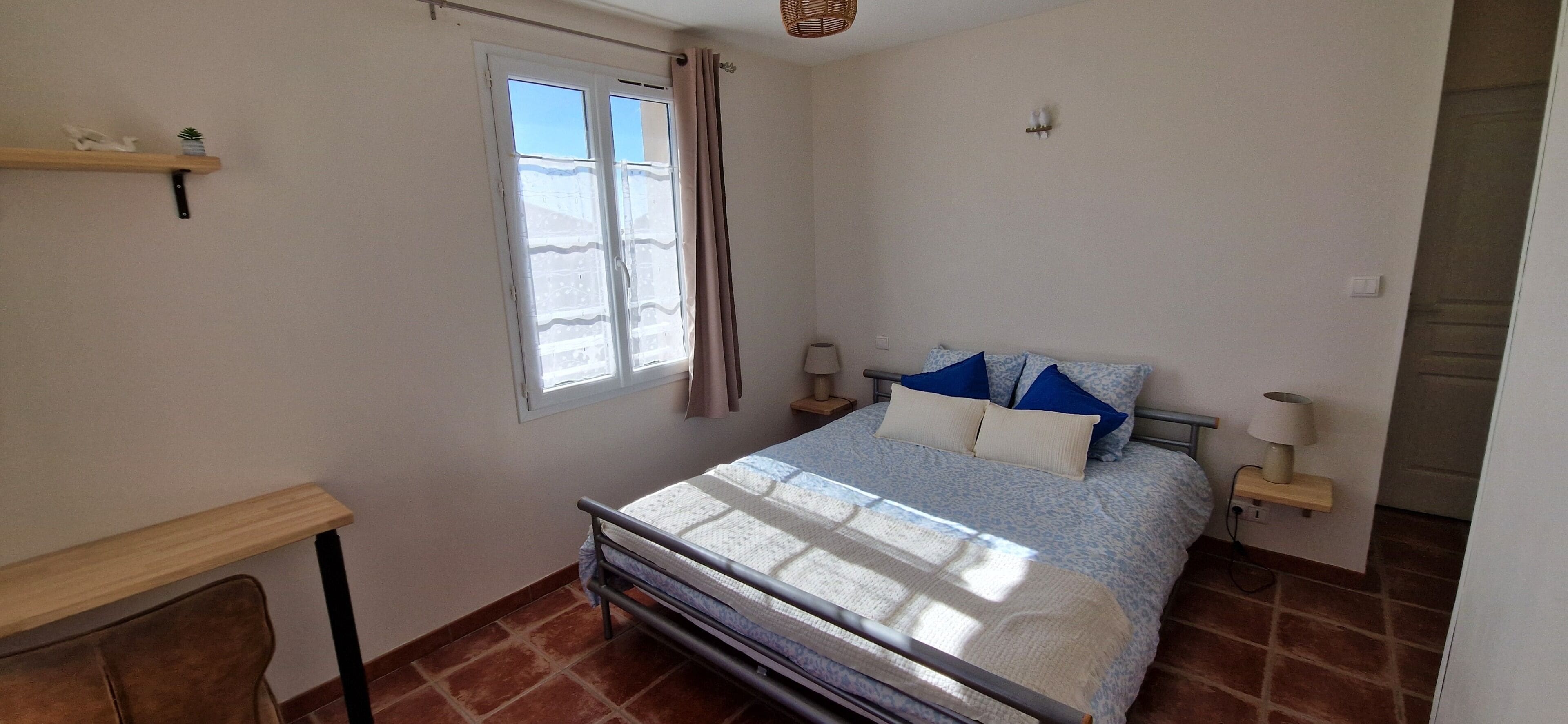 A bedroom with a bed, a window with curtains, a bedside table with a lamp, and a small shelf with decorative items.