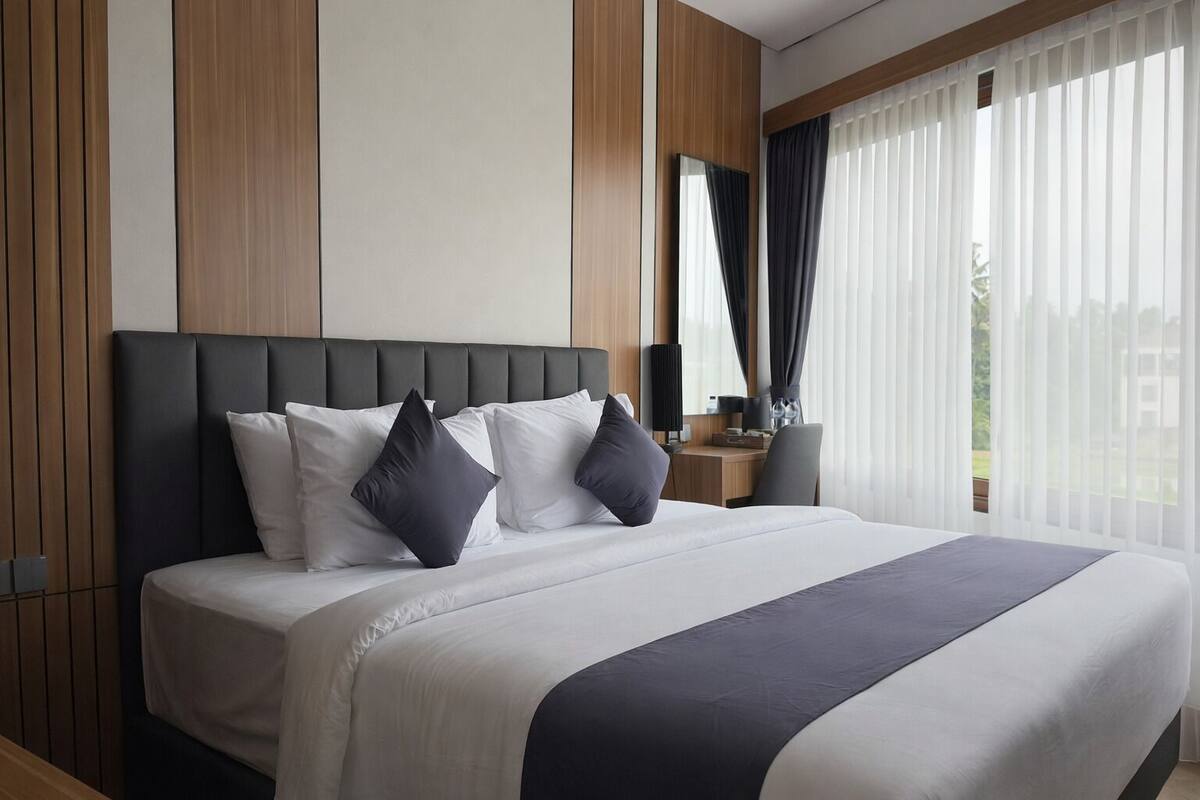 A modern hotel room with a large bed, a wooden headboard, a desk, and a large window with curtains.