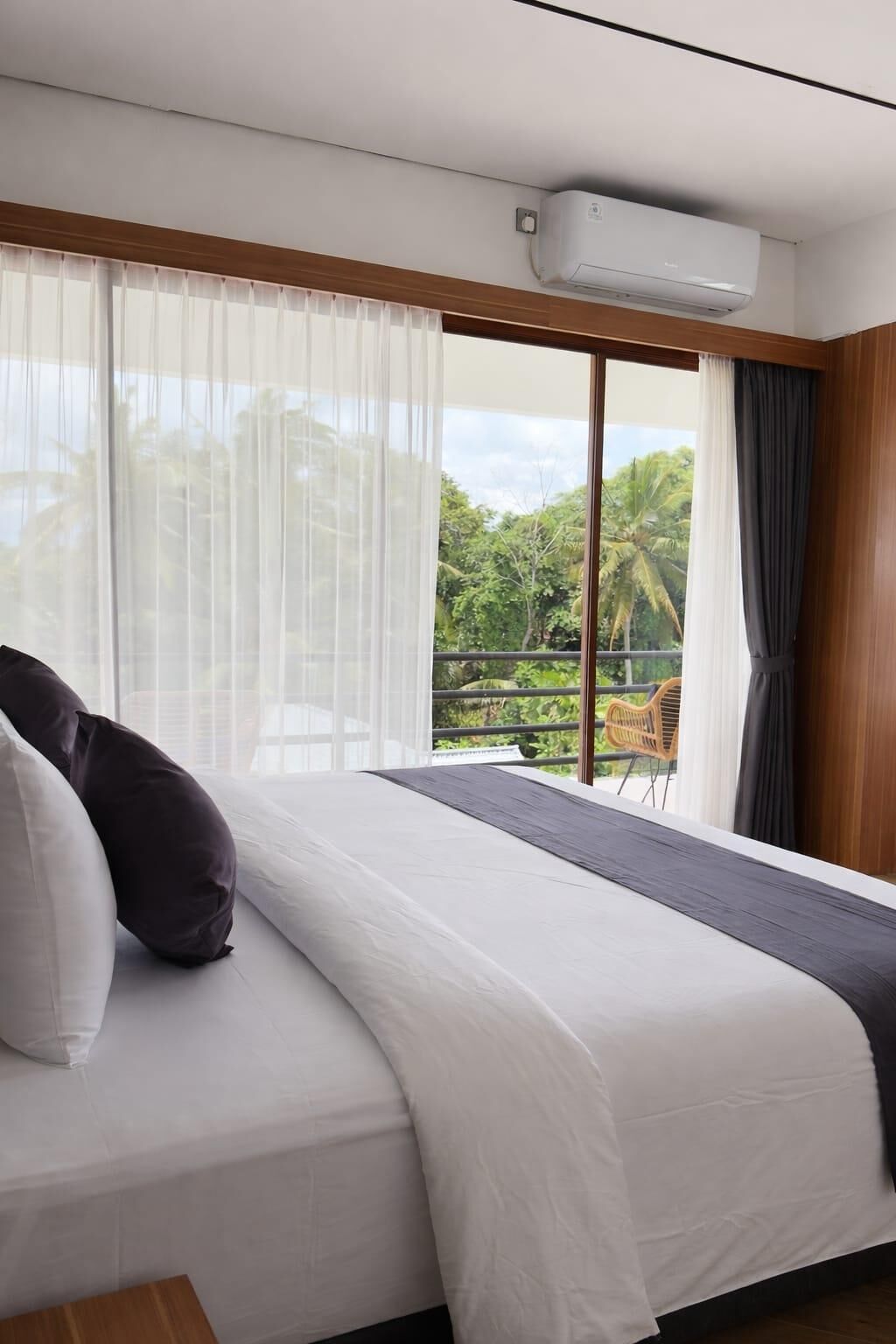 A hotel room with a bed, a window with sheer curtains, and a view of greenery outside.