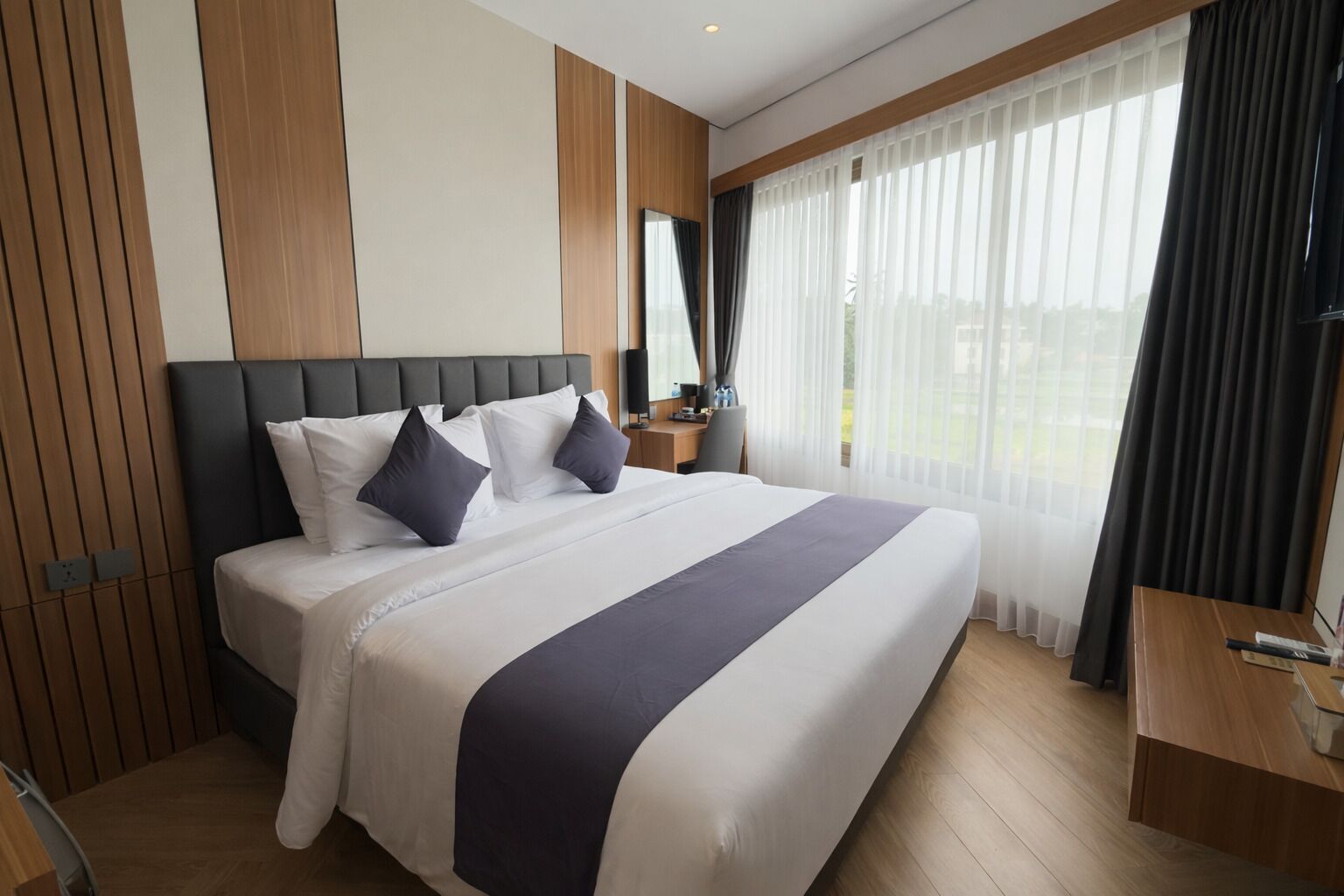 Habitación de hotel moderna con una cama grande, cabecera de madera, un escritorio y vistas al exterior a través de cortinas translúcidas.