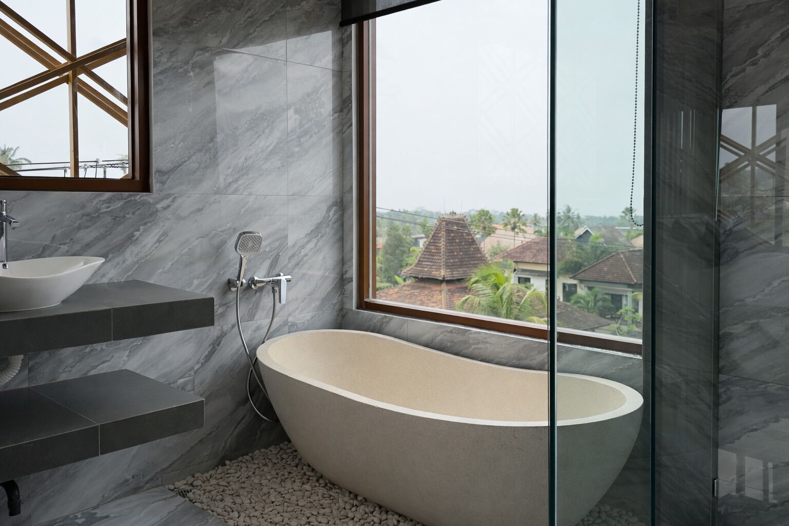 A modern bathroom with a freestanding bathtub, a large window offering a view of buildings, and a marble-tiled wall.
