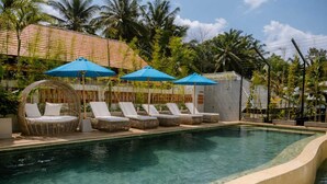 A pool area with lounge chairs and umbrellas, surrounded by palm trees and a building with a tiled roof. - Rendezvous Wellness & Resort (Ubud)