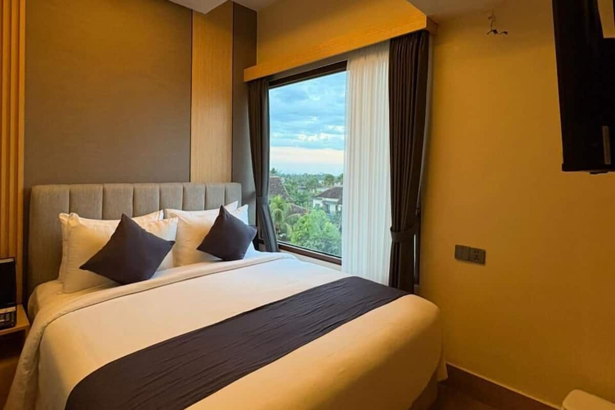 A hotel room with a bed, a window with curtains, an air conditioning unit, and a ceiling light fixture.