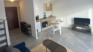 A modern living room with a blue sofa, a coffee table, and a TV on a wooden stand. There is a kitchen area with a microwave, sink, and cabinets.