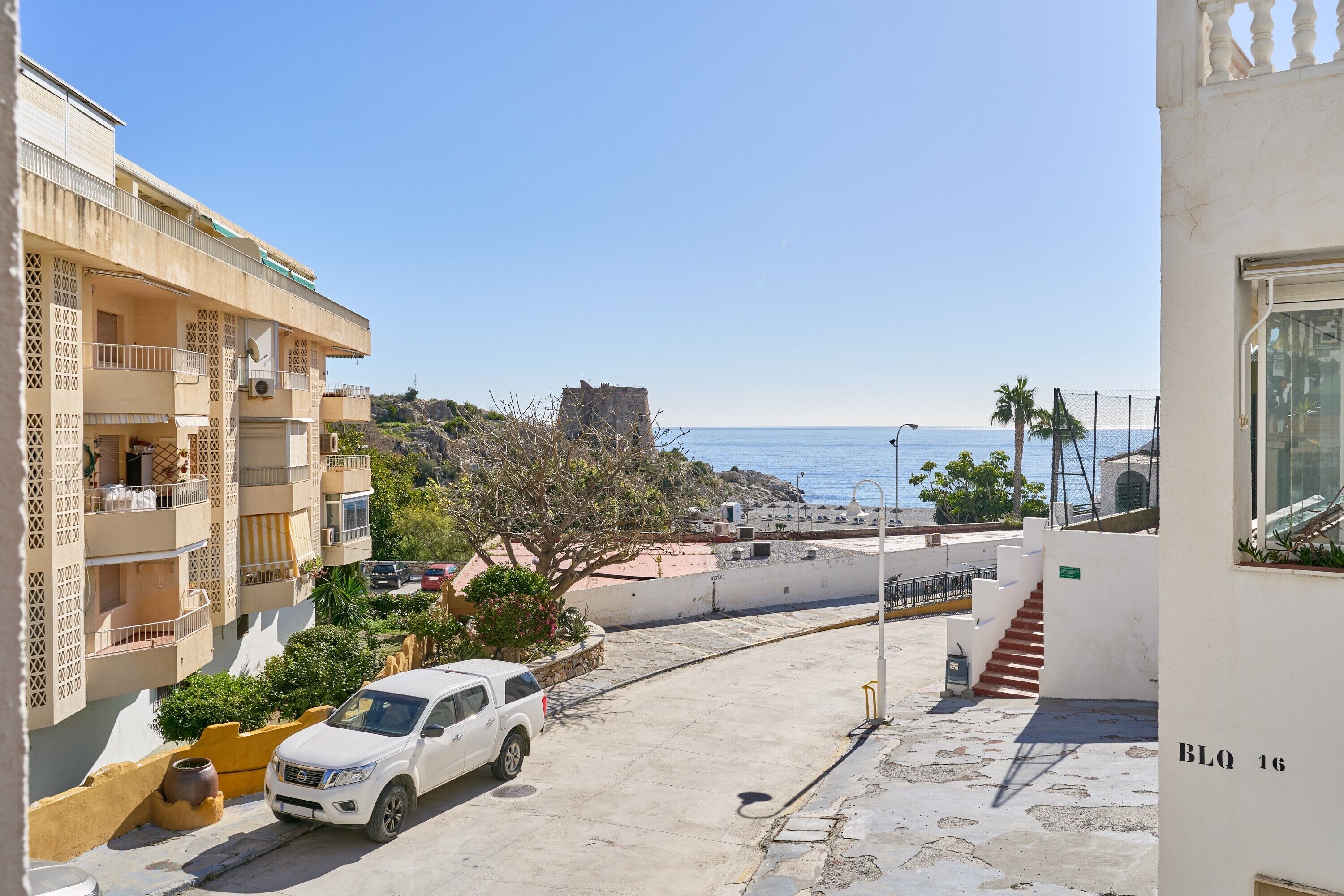 A residential building with balconies, a white car parked in front, and a view of the sea.