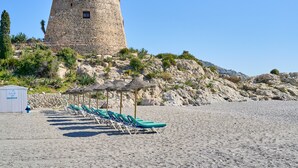 En strand med ett stentorn, solstolar och en liten byggnad.