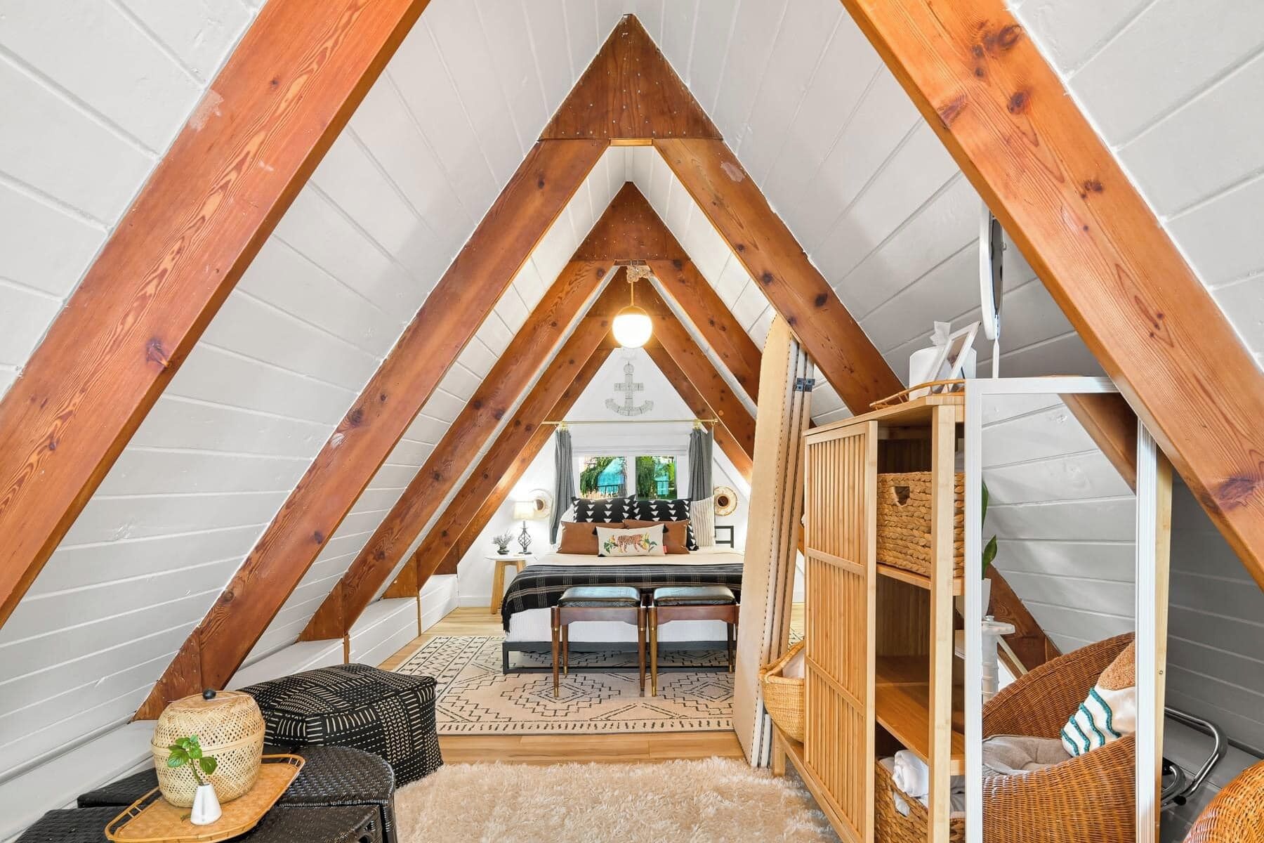 Une chambre mansardée chaleureuse avec un plafond en pente, des poutres en bois, un lit, une petite table et un panier en osier.