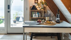 Una mesa de comedor con una botella de vino, un plato de frutas y un mantel a rayas. En el fondo, una estantería con libros y objetos decorativos.