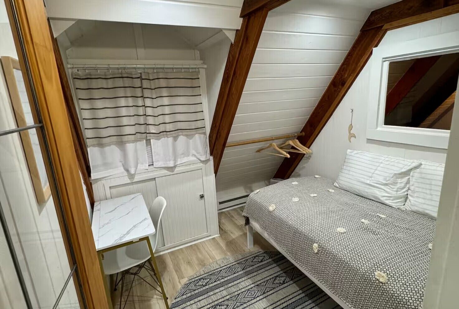 A small, compact room with a bed, a desk, and a chair. There is a window with a curtain, and the room has a sloped ceiling with wooden beams.