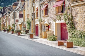 Une rangée de bâtiments en pierre avec des volets rouges, des plantes en pot et un banc sur le trottoir.