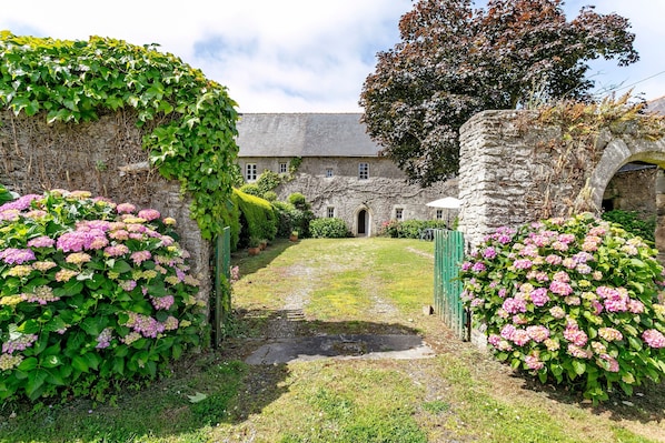 A stone building with a grassy courtyard, a stone archway, and flowering bushes.