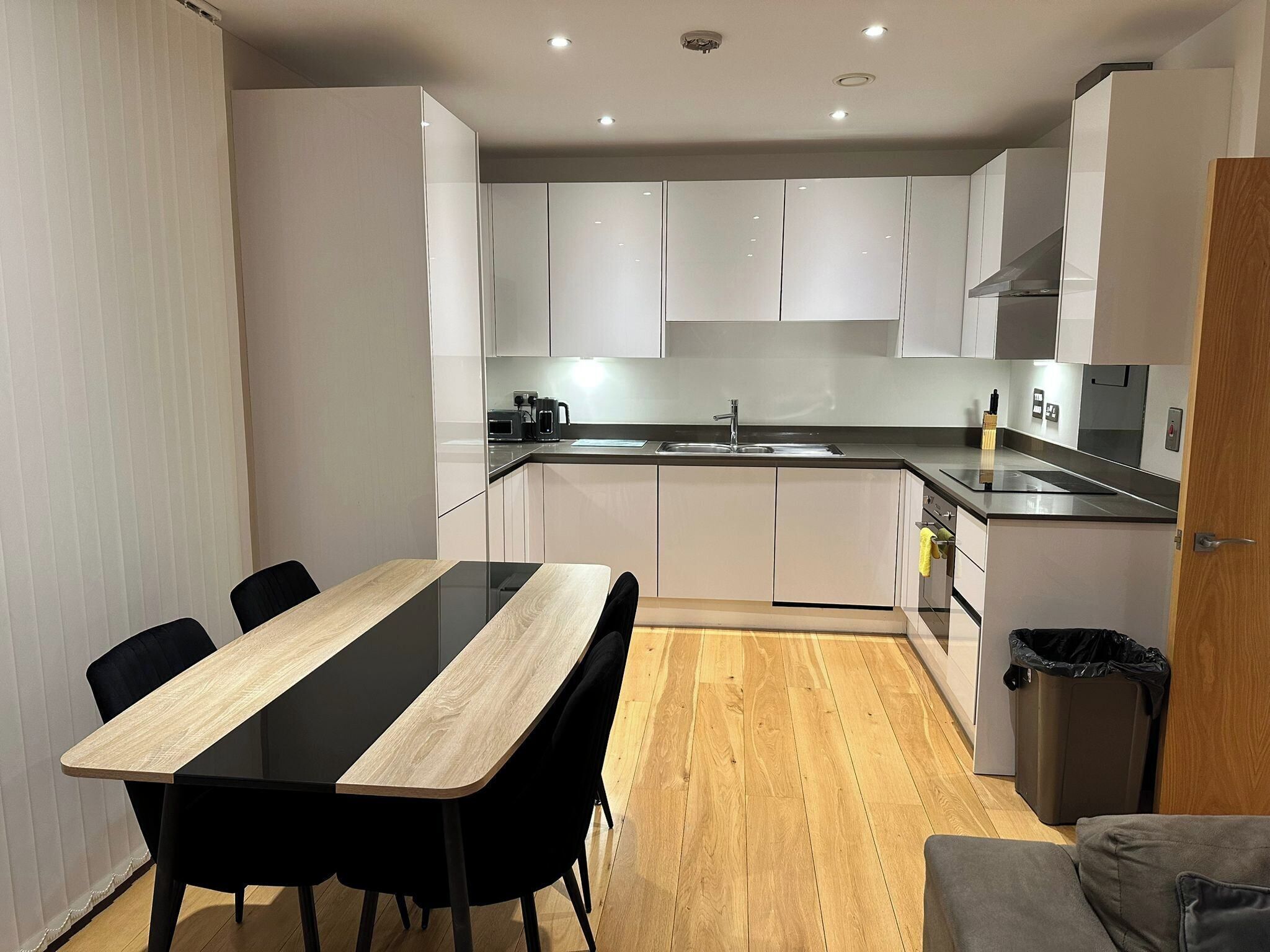 Eine moderne Küche mit weißen Schränken, einer schwarzen Arbeitsplatte, einem Spülbecken und einem Essbereich mit einem Holztisch und schwarzen Stühlen.