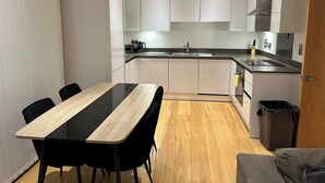 A modern kitchen with white cabinets, a black countertop, a sink, and a dining area with a wooden table and black chairs. - Luxury two bed two Bath In Excel (London)