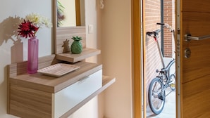 Een hal met een houten deur, een spiegel, een plank met een bloevas, en een fiets buiten.